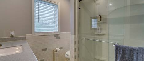 A bathroom with a glass-enclosed shower, a sink, a toilet, and a window with blinds.