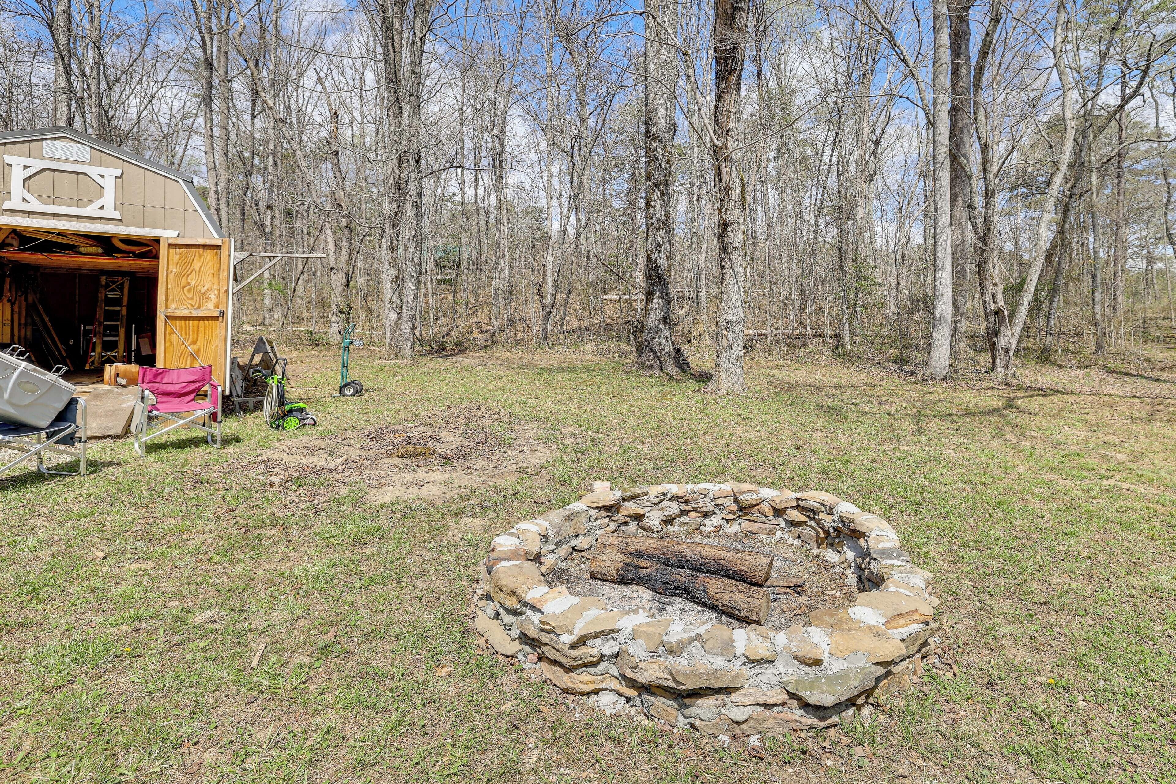Une cabane rustique avec un foyer en pierre dans la cour, entourée d’arbres.