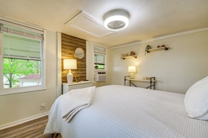 A bedroom with a bed, bedside table, lamp, mirror, and a window with blinds.