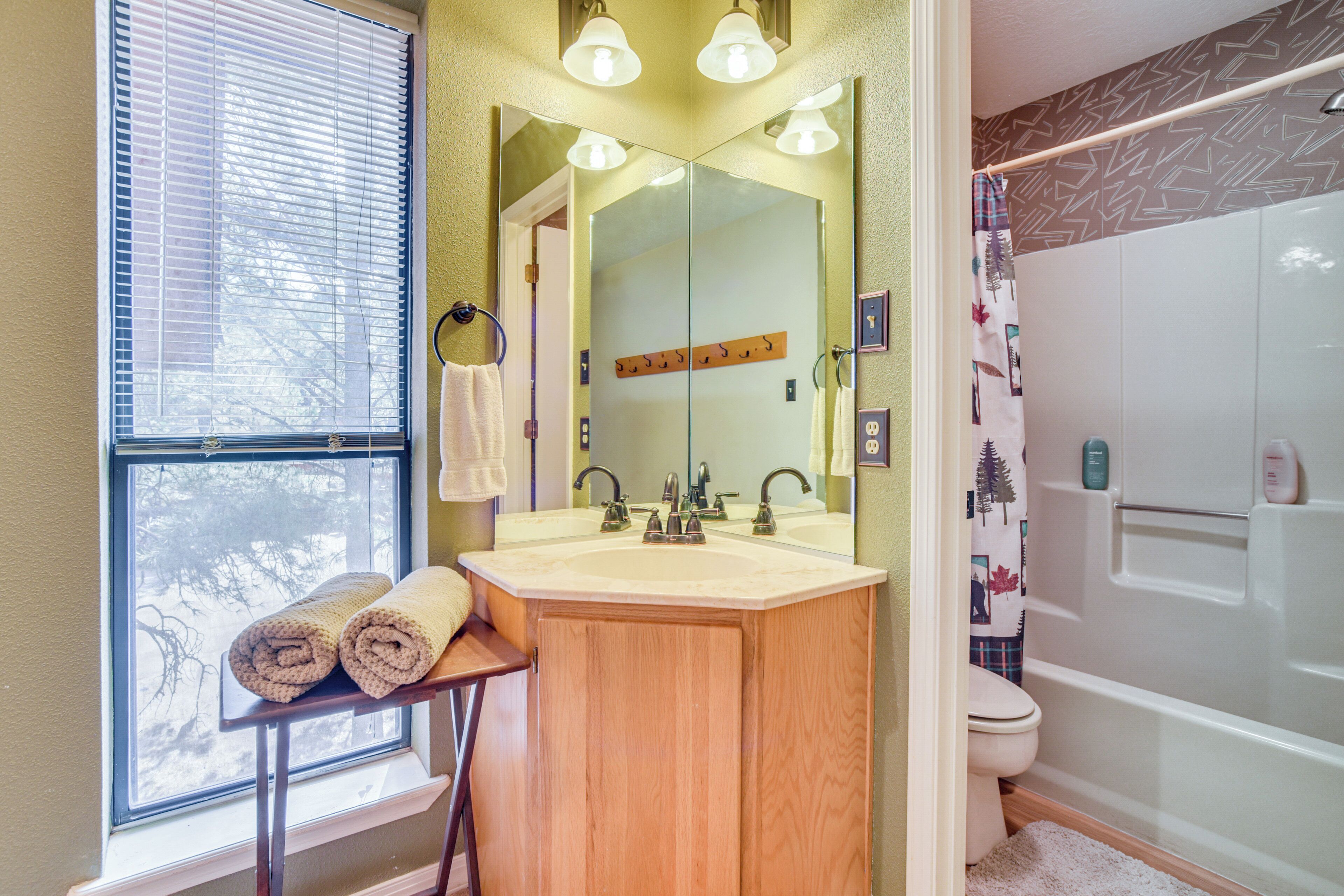 A bathroom with a wooden vanity, a large mirror, a window with blinds, a shower, and a toilet.