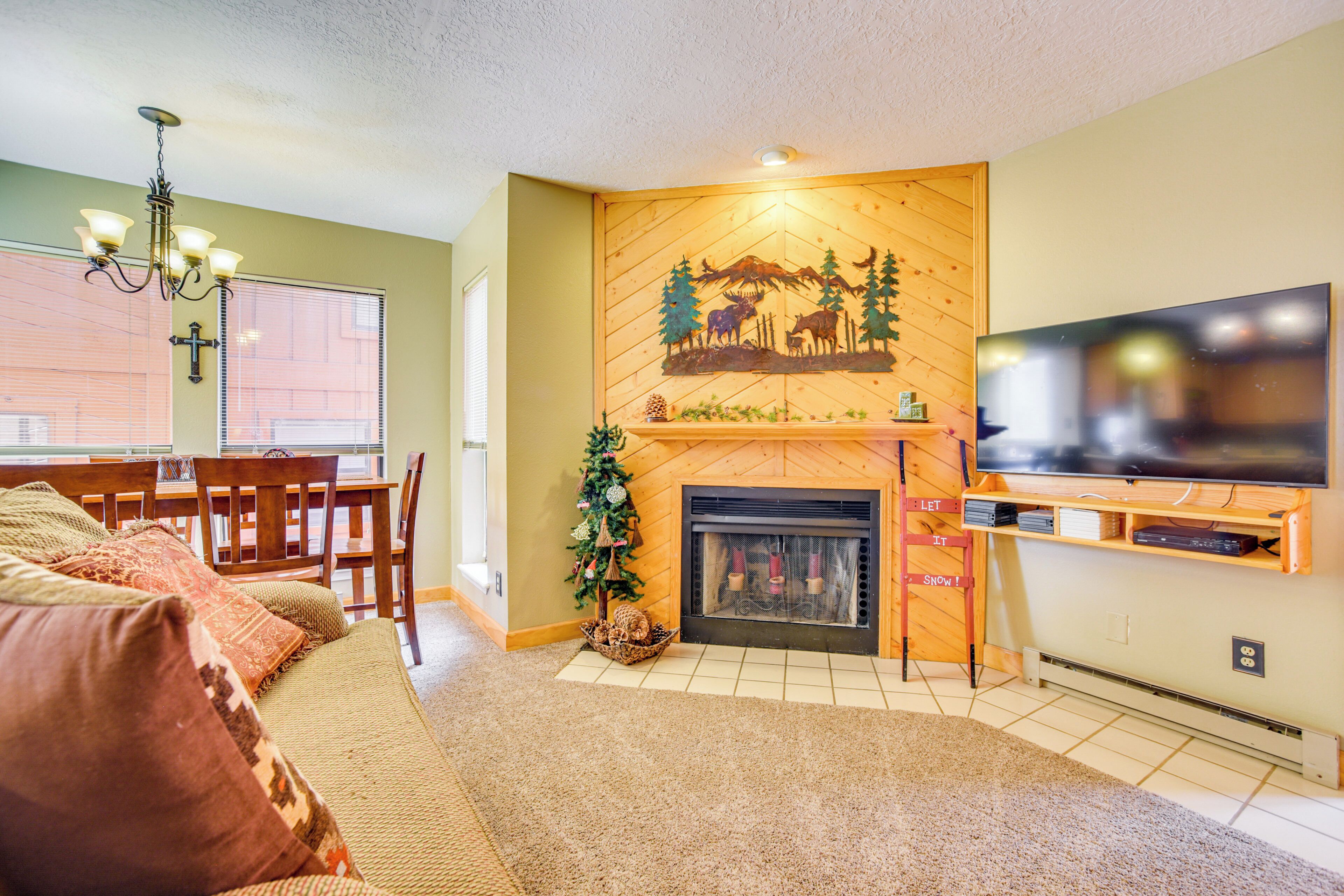 A living room with a fireplace, a TV mounted on the wall, a dining area with chairs, and a Christmas tree.