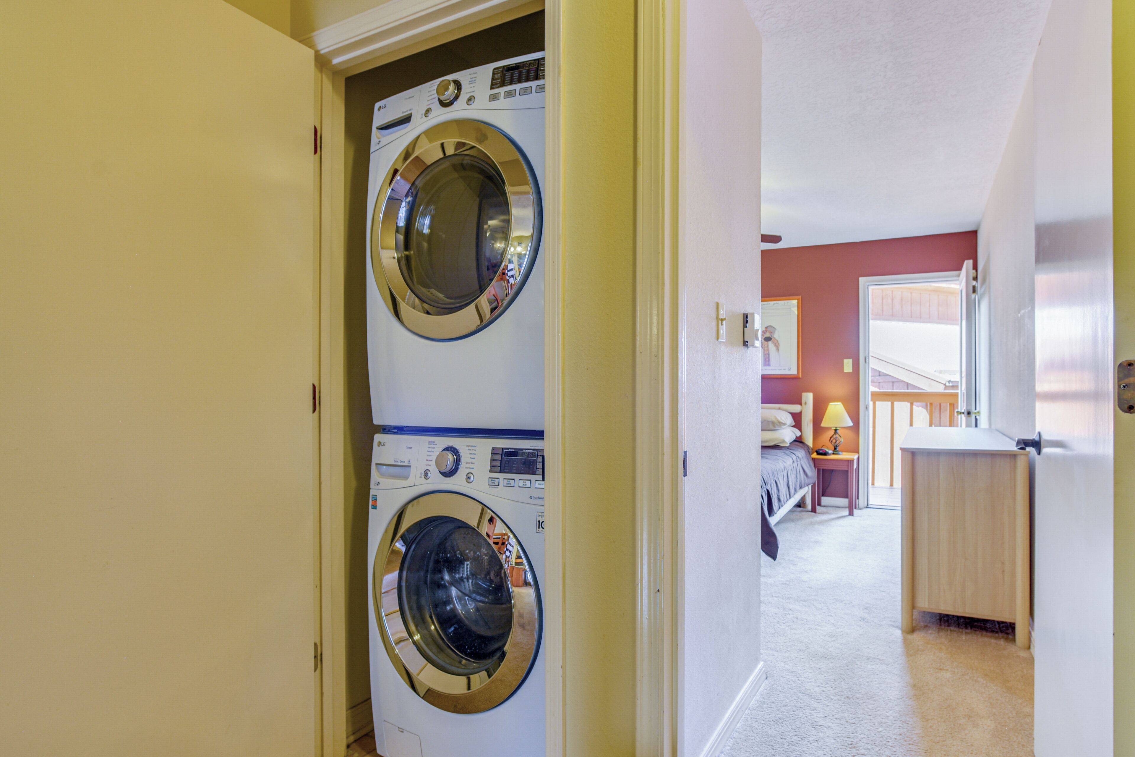 A laundry room with two front-loading washing machines and a bed in the adjacent room.