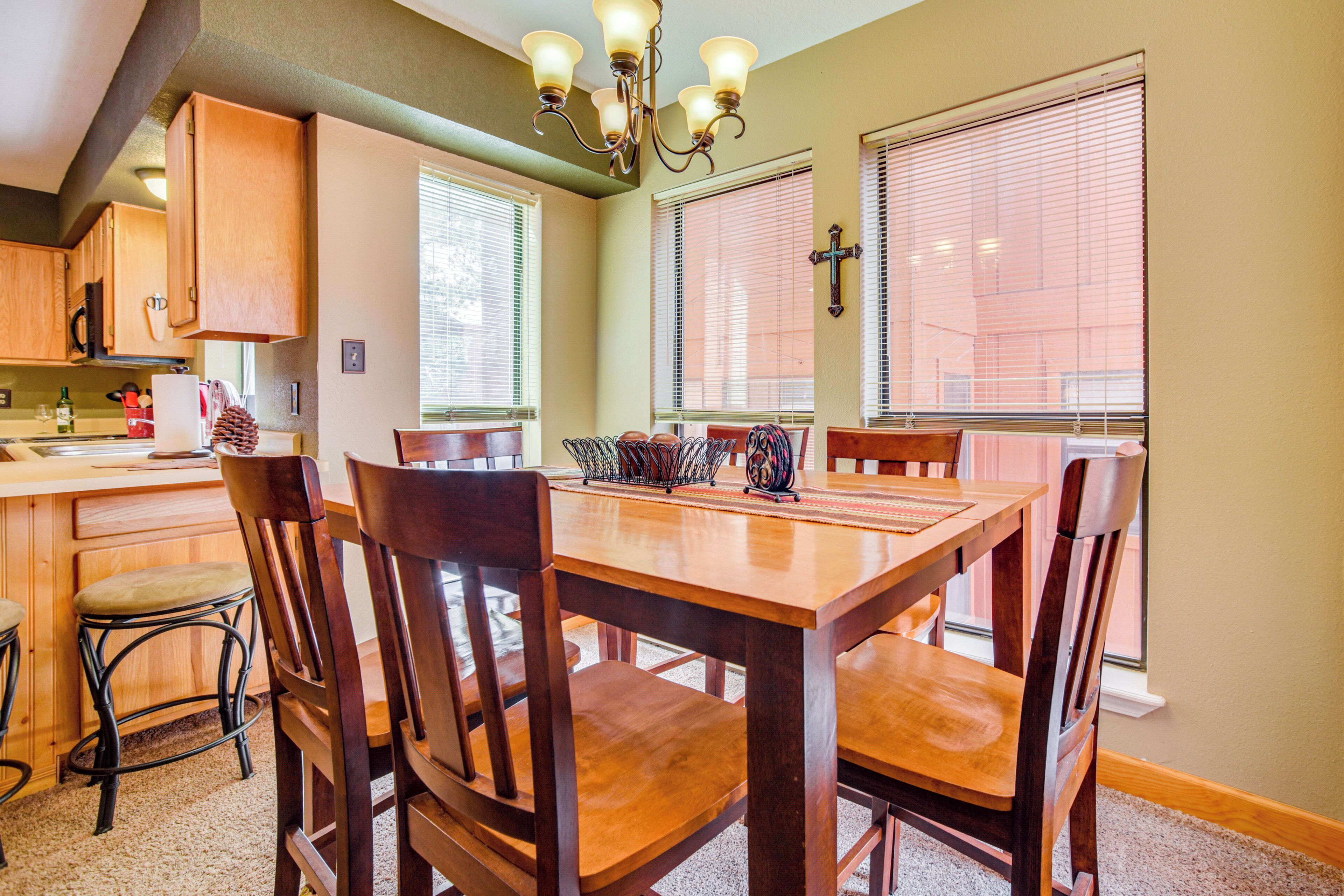 A dining area with a wooden table and chairs, a chandelier, and a kitchen in the background.