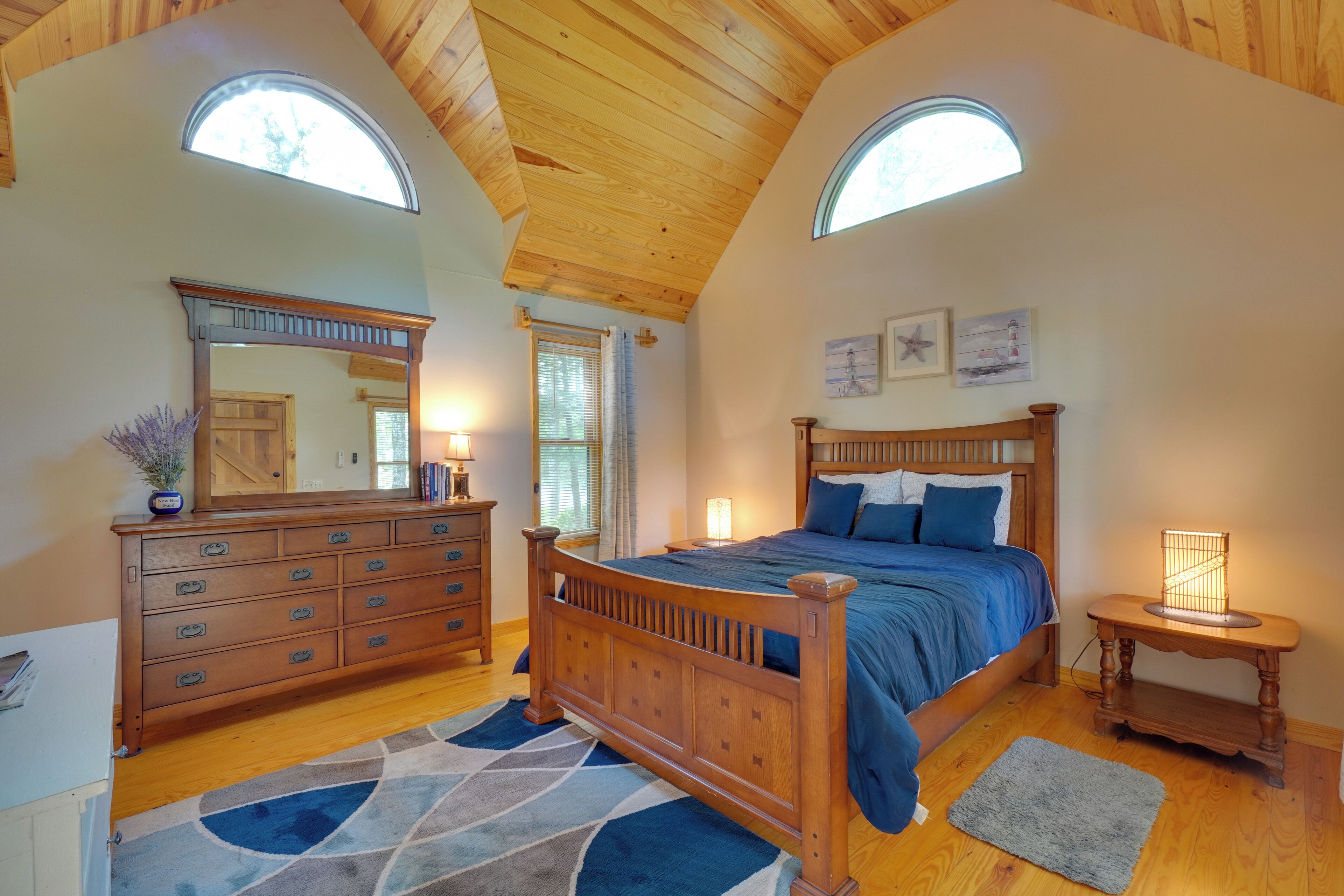 A bedroom with a wooden ceiling, a bed with blue bedding, a dresser, a mirror, and a nightstand with a lamp.