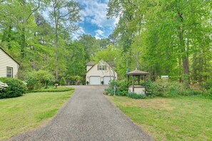 Une allée de gravier mène à une maison dotée d’un garage, le tout étant entouré d’une végétation luxuriante et d’arbres.