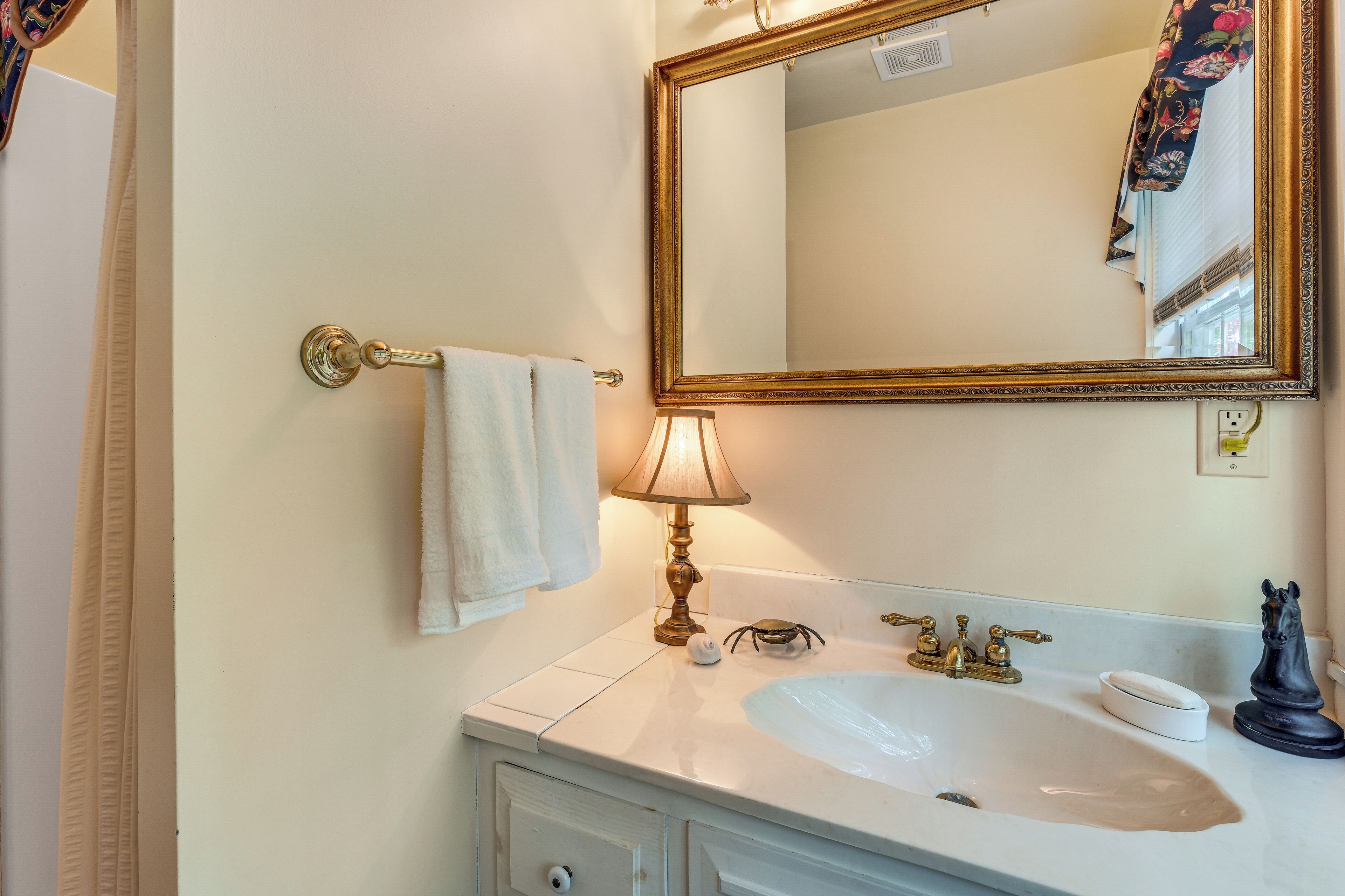 A bathroom with a white sink, gold faucets, a mirror with a gold frame, a towel hanging on a towel rack, and a lamp.