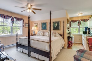 A four-poster bed with a ceiling fan, a window with plaid curtains, and a kitchen area with a sink and cabinets.