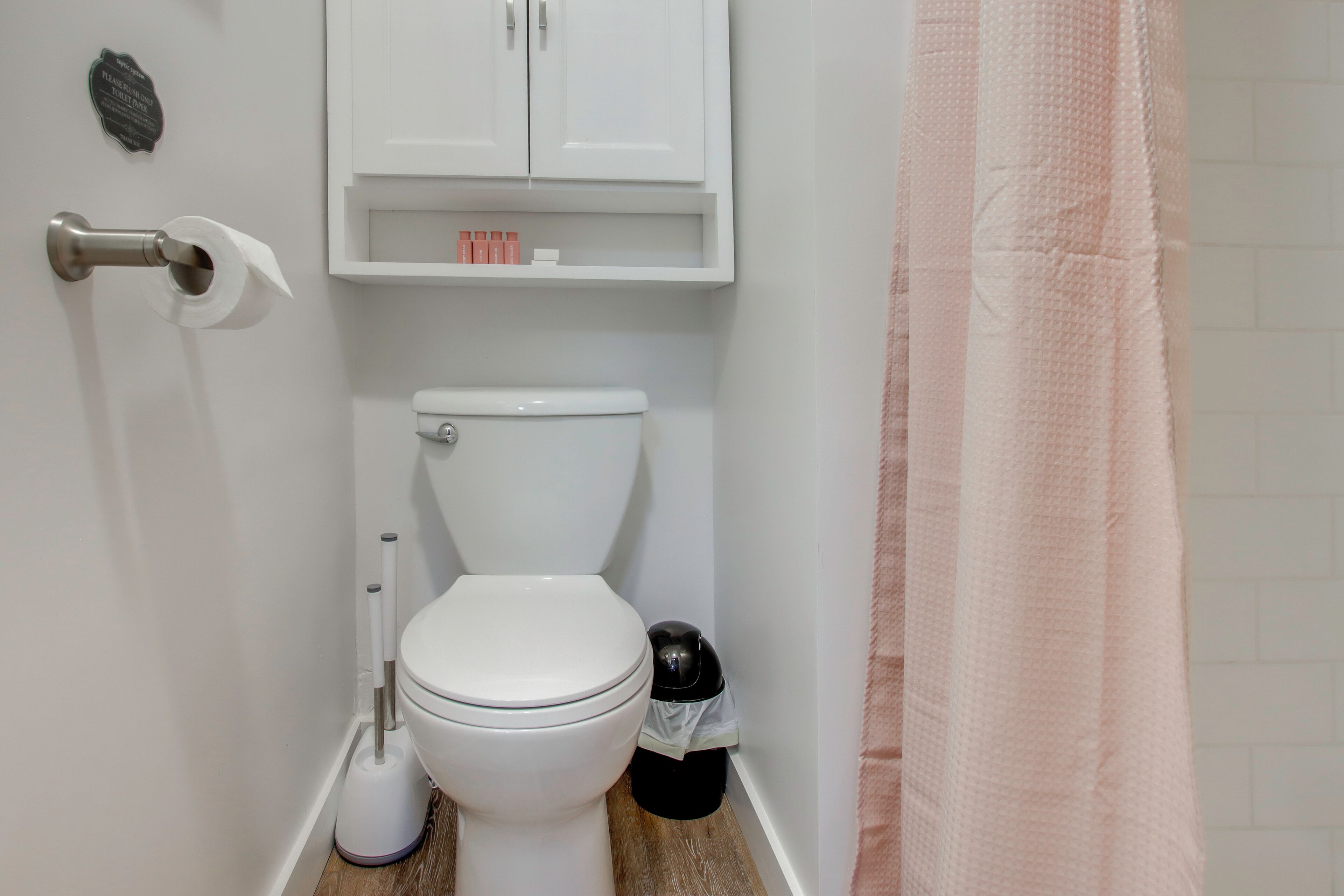 Een badkamer met een toilet, een wit kastje, een prullenbak en een roze douchegordijn.