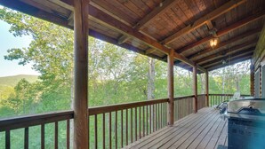 A covered outdoor deck with a grill, wooden railings, and a view of a forested area. - Wooded Cabin w/ Decks & Dale Hollow Lake Views! (Byrdstown)