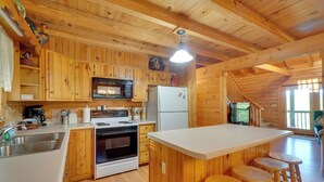A wooden cabin kitchen with a central island, bar stools, a microwave, stove, refrigerator, and a staircase leading to a deck. - Wooded Cabin w/ Decks & Dale Hollow Lake Views! (Byrdstown)