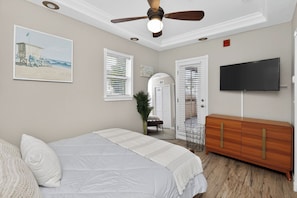 A bedroom with a bed, a ceiling fan, a TV, a dresser, and a beach-themed painting.