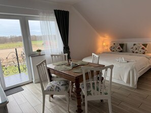 Ein Schlafzimmer mit einem großen Bett, einem Esstisch mit Stühlen, Blick durch ein Fenster und einem kleinen Balkon.