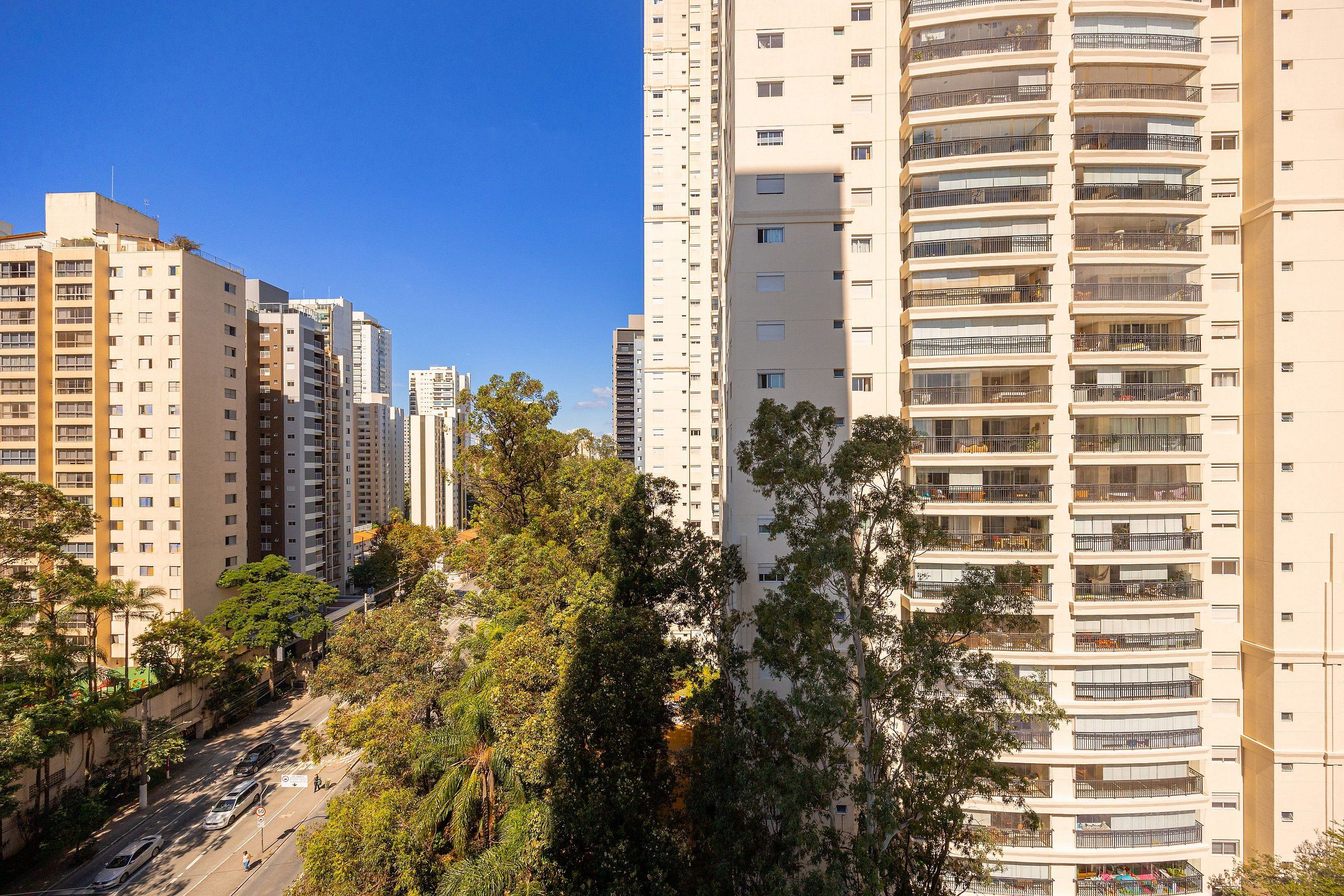 A high-rise residential building with multiple balconies, a clear sky, and surrounding greenery.