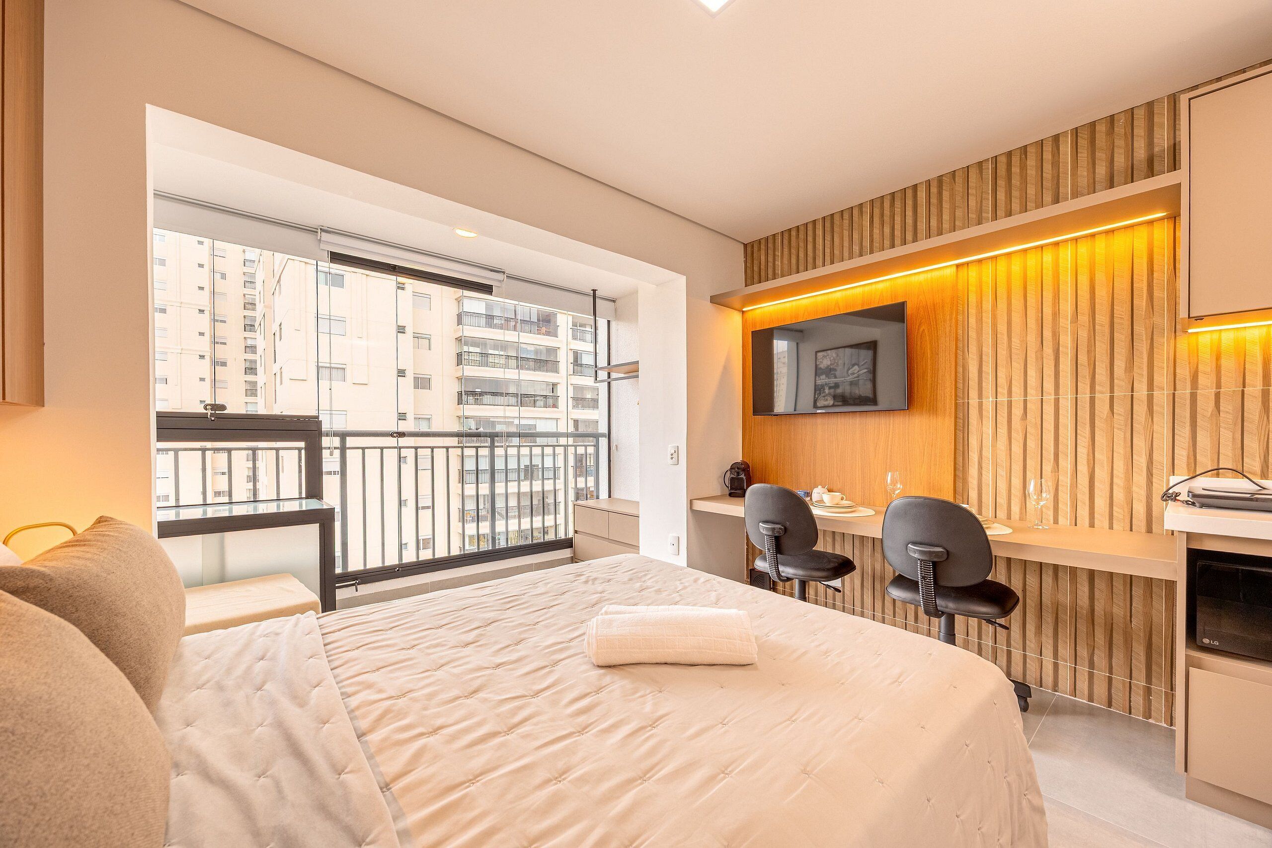 Quarto de hotel moderno com uma cama grande, uma escrivaninha com duas cadeiras, televisão e varanda com vista para a cidade.