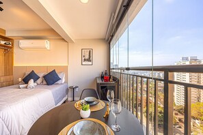 A hotel room with a bed, a small table with a vase and wine glass, and a balcony with a city view. - Hospedez - Estudios Campo Belo Movi (São Paulo)