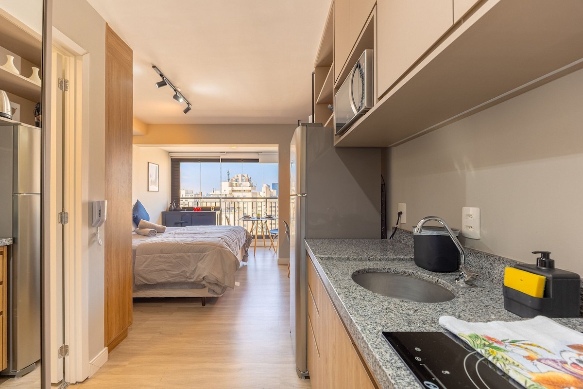 A compact hotel room with a kitchenette, a bed, and a view of the cityscape.