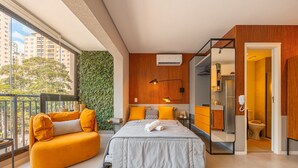 A modern bedroom with a bed, a sofa, a bathroom, and a view of the city. - Hospedez - Estudios Campo Belo Movi (São Paulo)