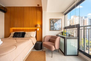 Ein modernes Schlafzimmer mit einem Bett, einem kleinen Hocker, einem Stuhl, einem Fenster mit Blick auf Gebäude und einem Bild an der Wand.