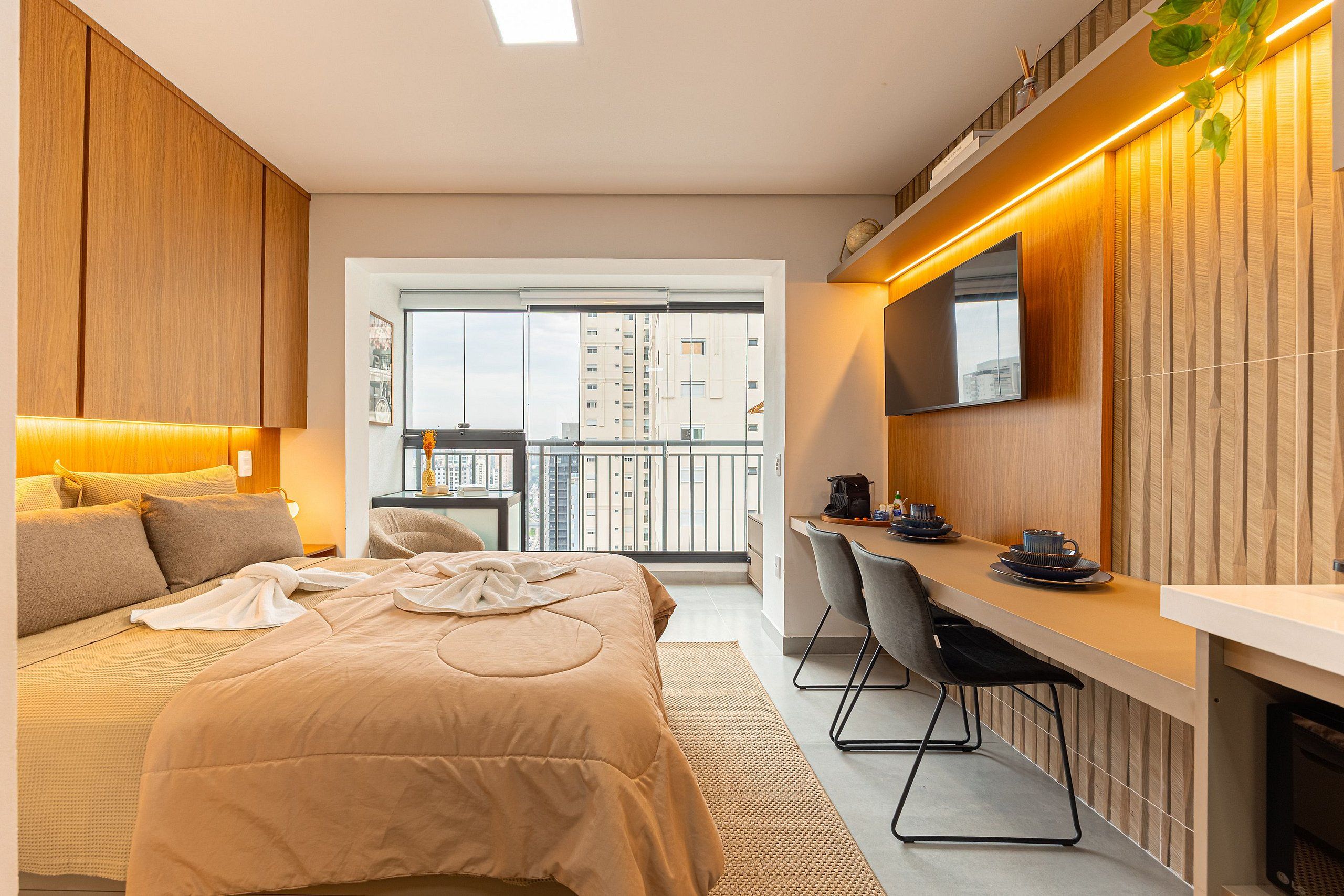 A modern hotel room with a bed, a desk, and a balcony with a city view.