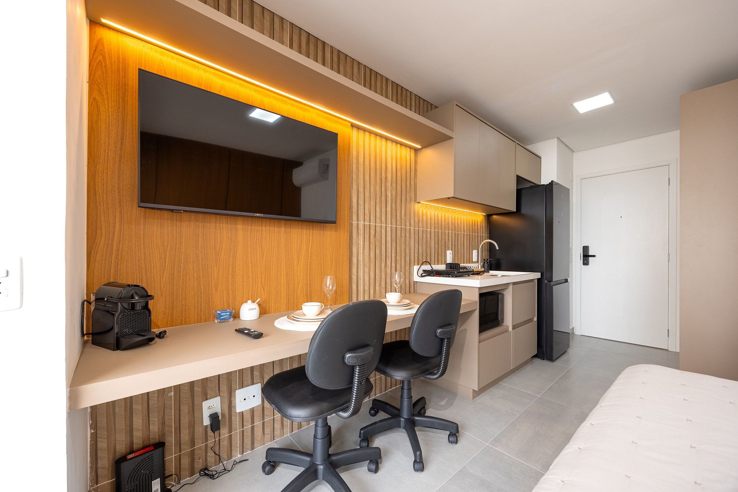 A modern kitchen with a built-in stove, a microwave, and a refrigerator. There are two black office chairs and a small table with a laptop and a cup.