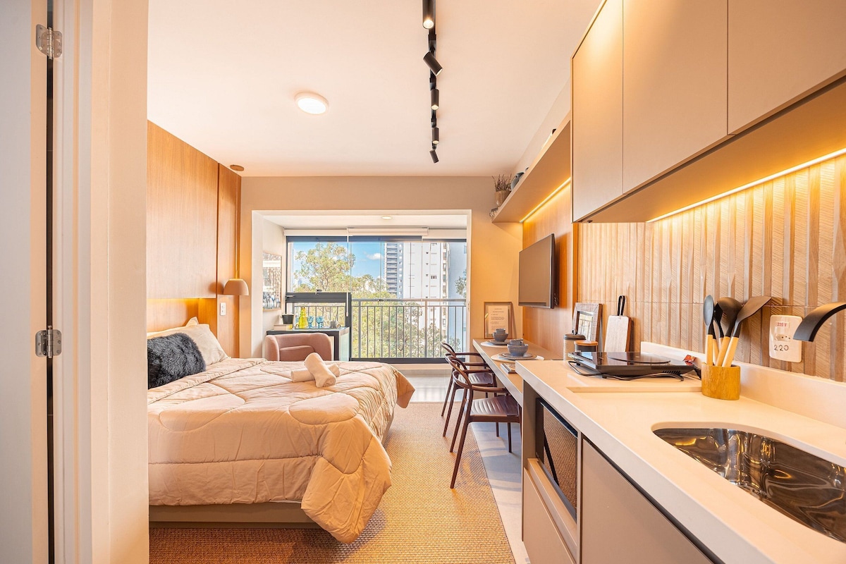 A modern hotel room with a bed, kitchenette, and a balcony with a city view.