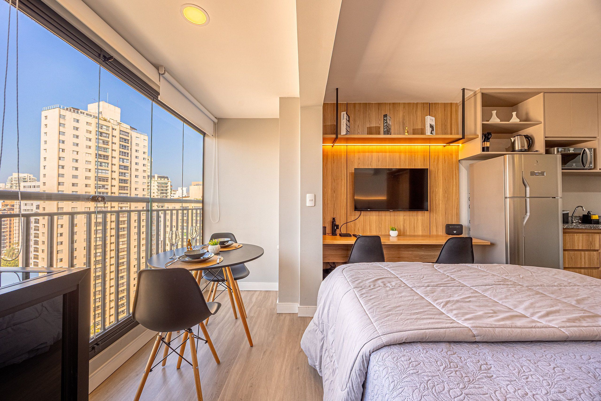 A modern hotel room with a balcony, a dining area, a kitchenette, and a bed.
