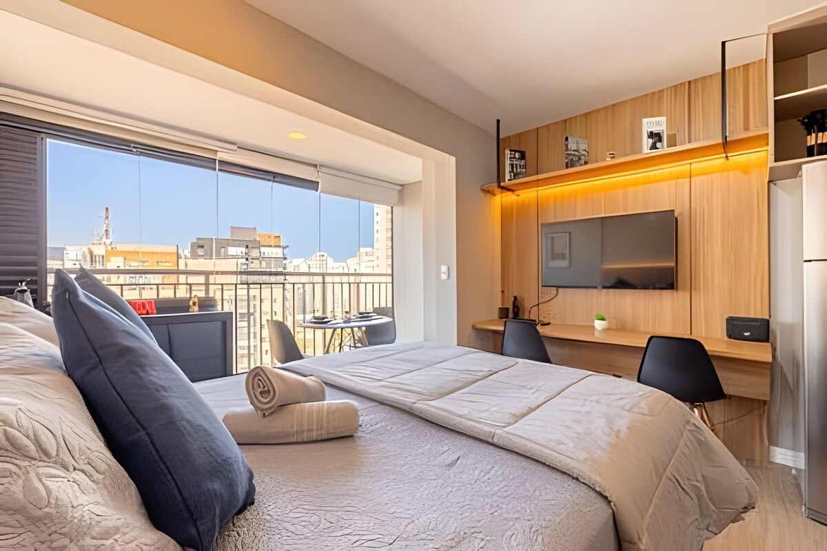 A modern hotel room with a large bed, a balcony with a city view, a flat-screen TV mounted on the wall, and a small table with chairs.