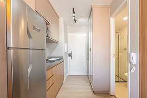 A narrow kitchen with a stainless steel refrigerator, wooden cabinets, and a white door at the end. - Hospedez - Estudios Campo Belo Movi (São Paulo)