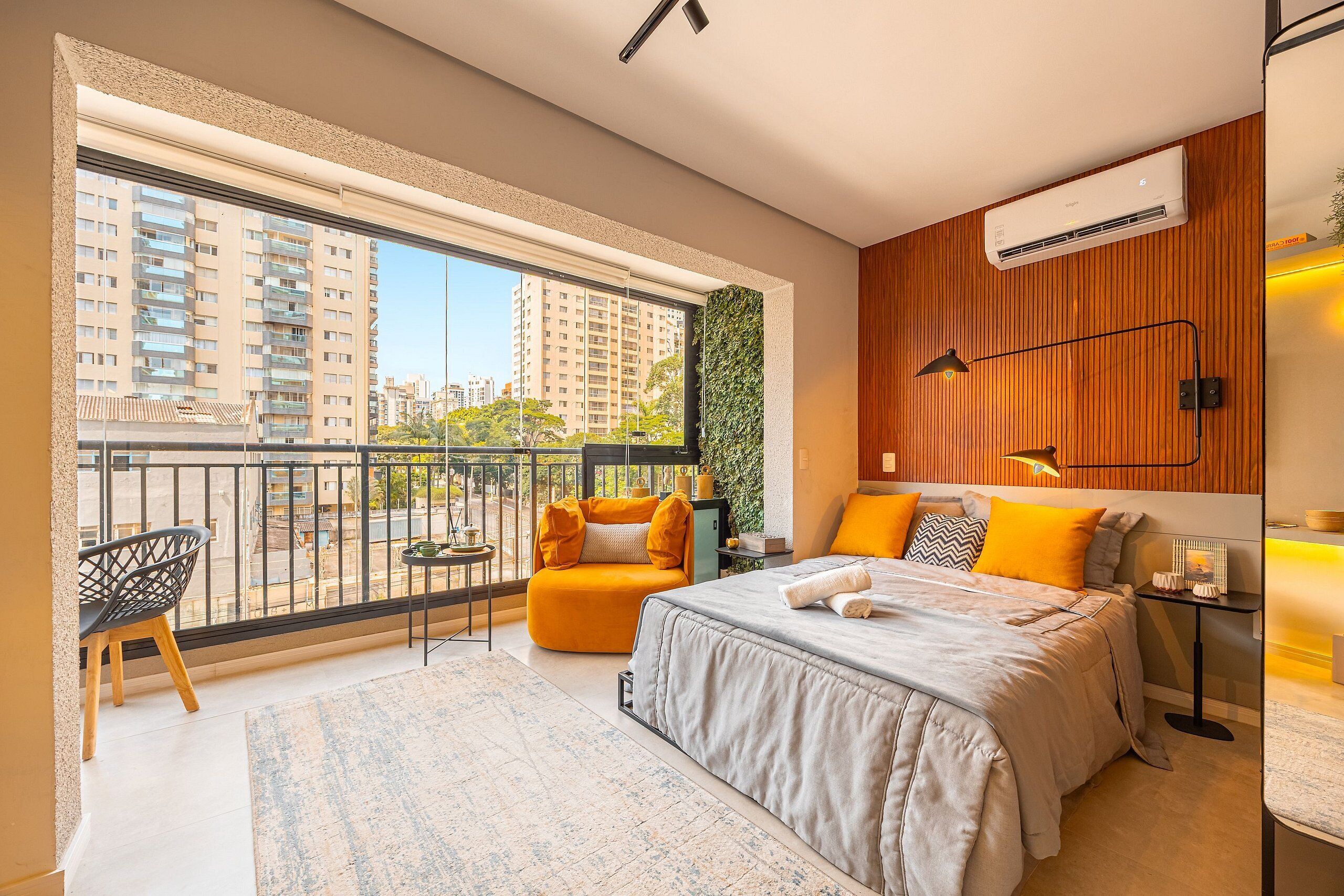 A modern bedroom with a large window, a balcony, a bed with a grey duvet, and a small table with a lamp.