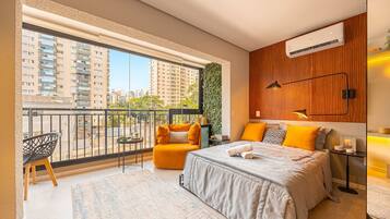 A modern bedroom with a large window, a balcony, a bed with a grey duvet, and a small table with a lamp.