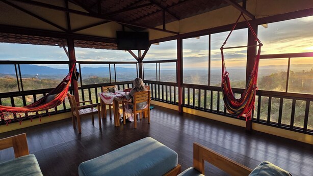 Una habitación con suelo de madera, una mesa con sillas, una hamaca y un ventanal con vistas al paisaje.