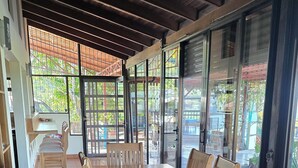 Un espacio cubierto para comer con una gran mesa de madera y sillas, ventanas amplias y vistas al exterior.