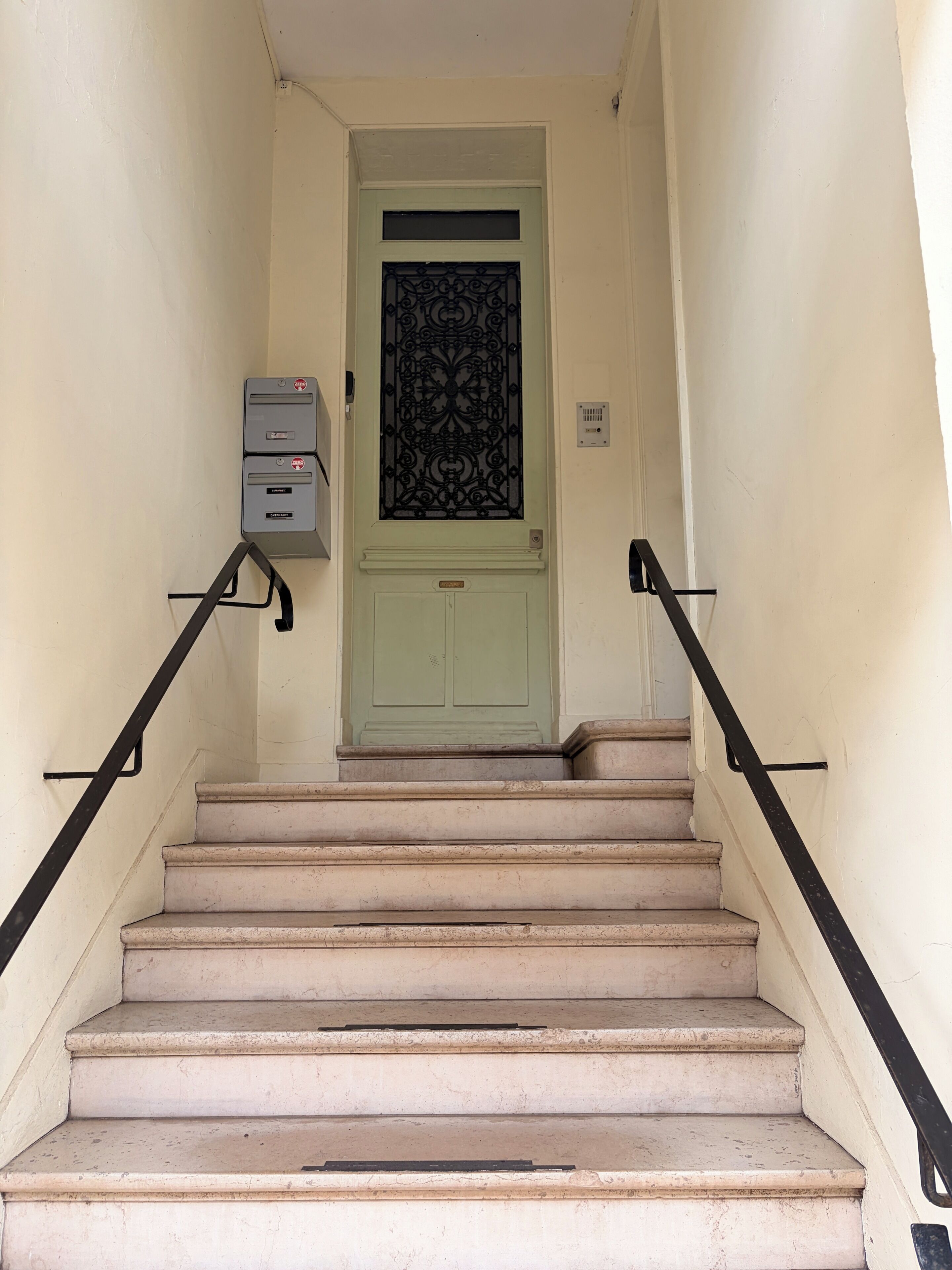 Un escalier avec une porte vert clair, une boîte aux lettres et une rampe métallique.