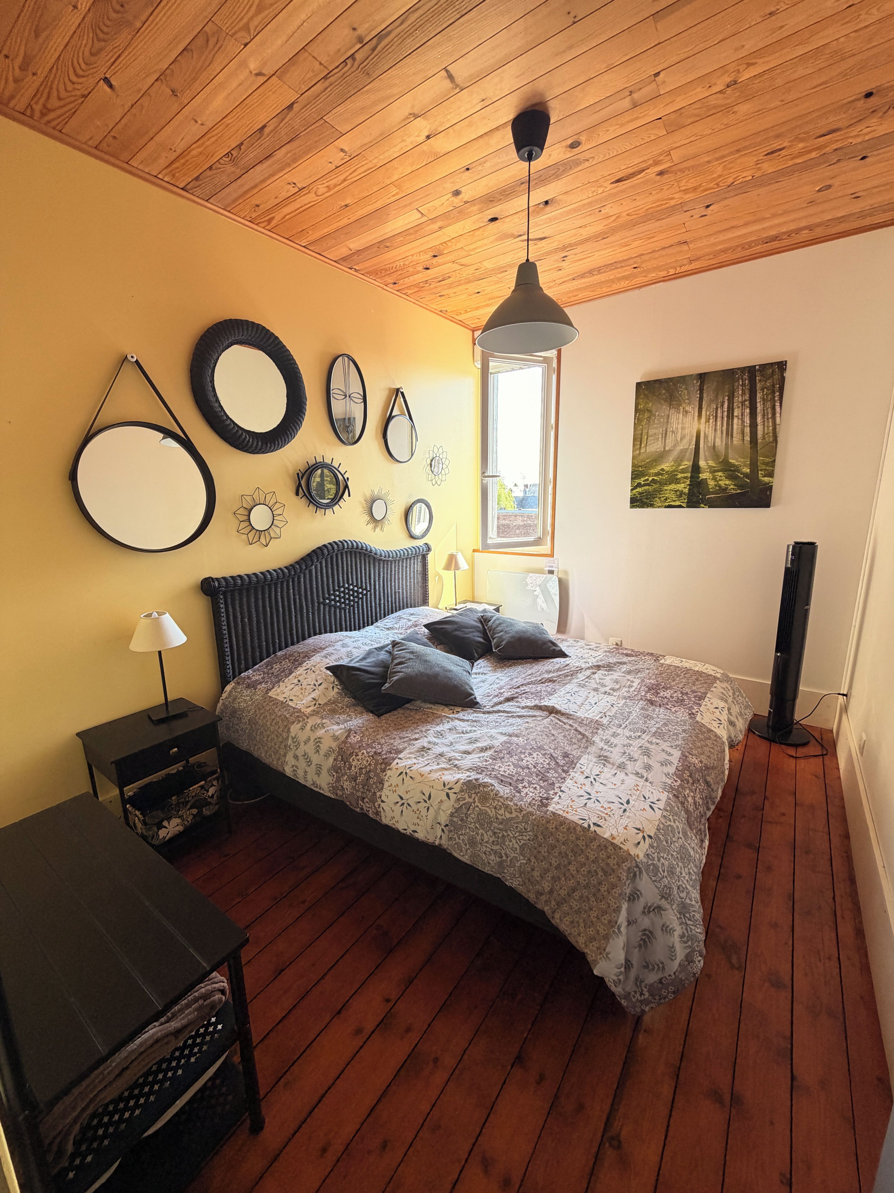 Ein Schlafzimmer mit Holzboden, einem Bett mit gemustertem Bettbezug, einem Nachttisch mit Lampe und Wanddekorationen, darunter Spiegel und ein Waldgemälde.