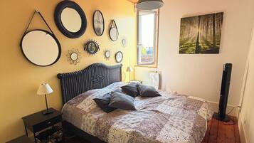 Ein Schlafzimmer mit Holzboden, einem Bett mit gemustertem Bettbezug, einem Nachttisch mit Lampe und Wanddekorationen, darunter Spiegel und ein Waldgemälde.
