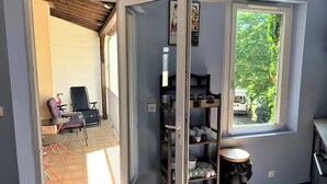 A room with a sliding door leading to an outdoor seating area, a window with a view of greenery, and a built-in shelf with kitchenware.