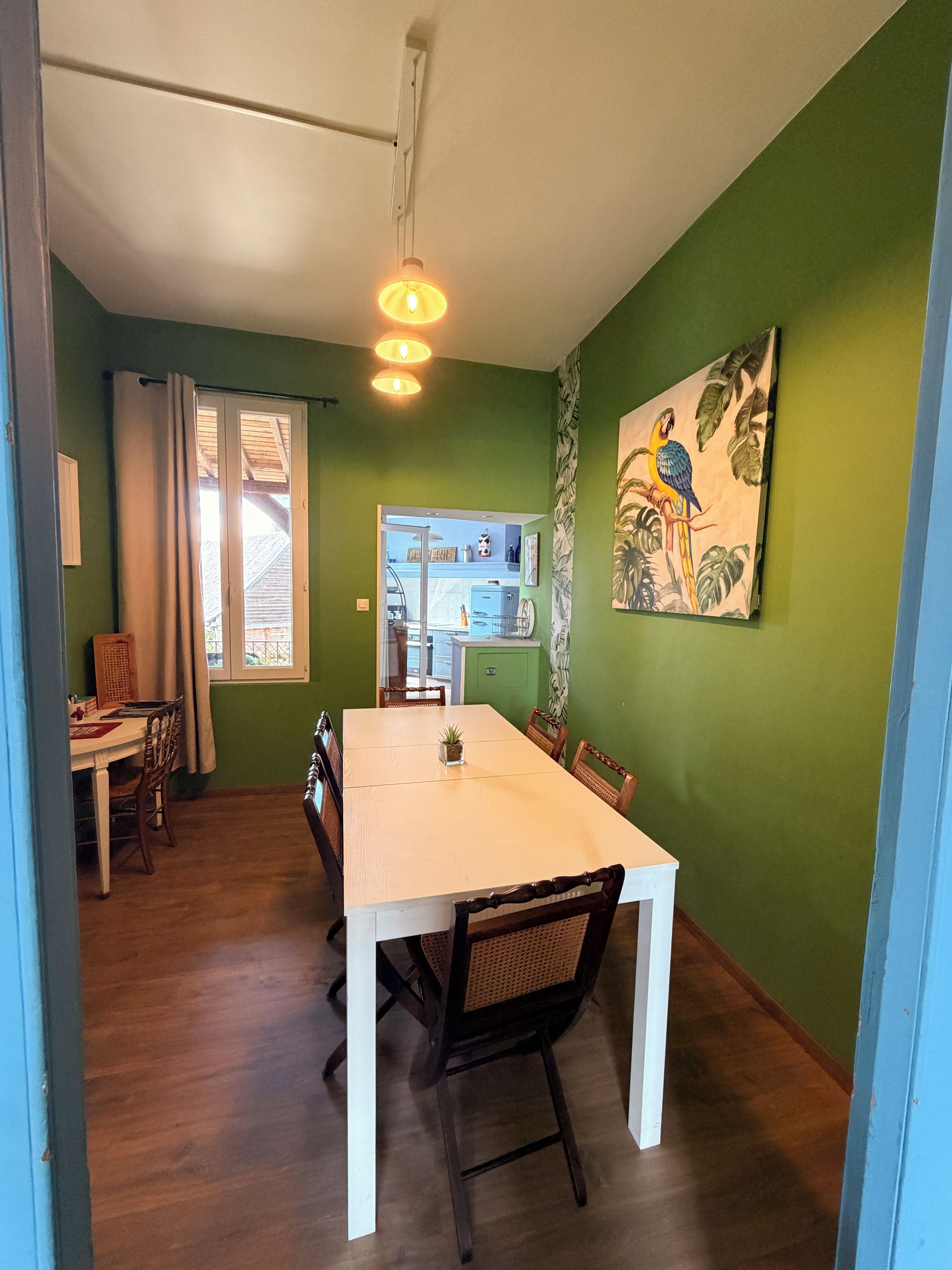 Un comedor con mesa y sillas blancas, una pared verde con un cuadro de un loro, y vista a la cocina.