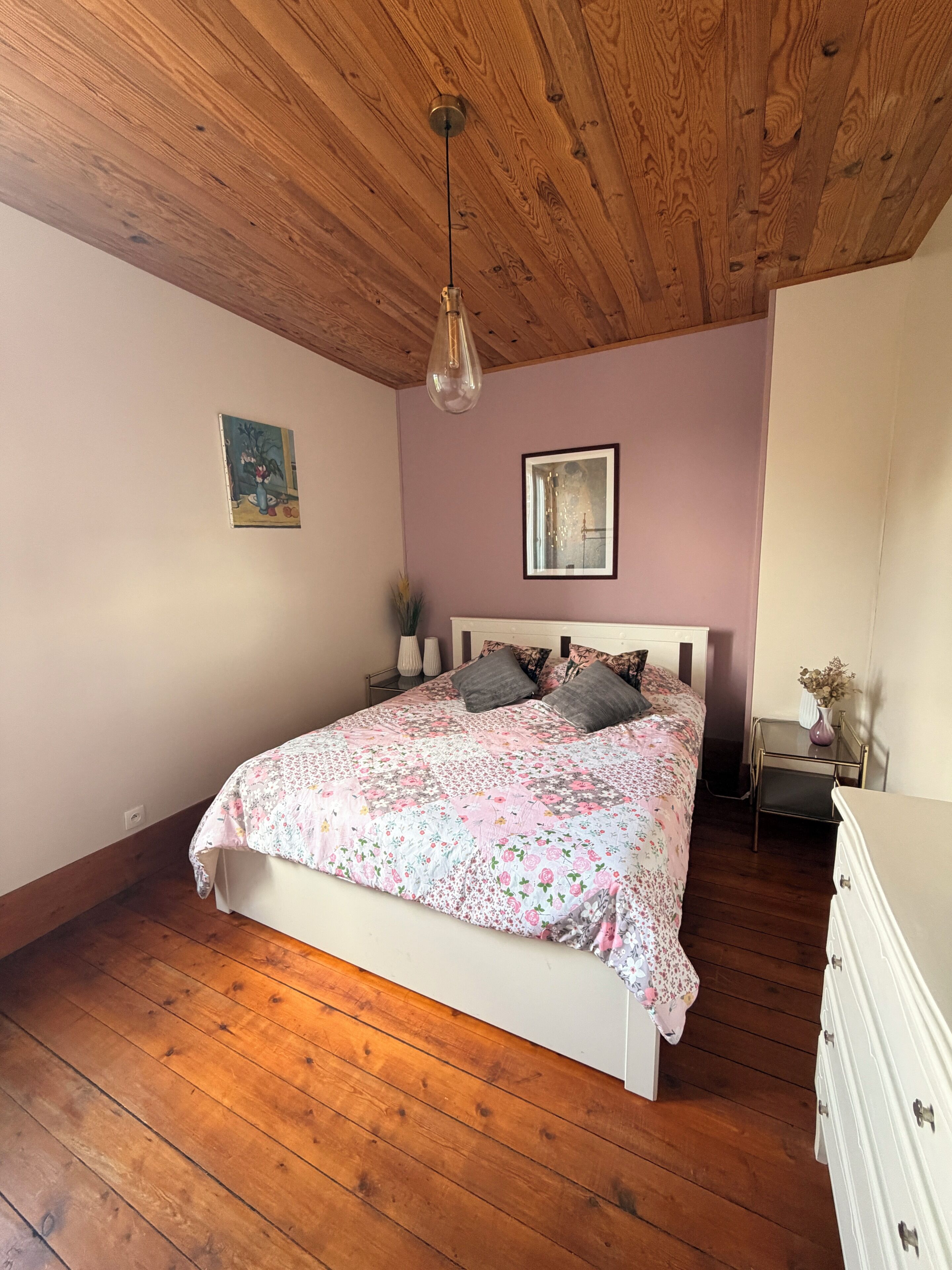 Una camera da letto con soffitto in legno, un letto con biancheria da letto floreale, un comodino con un vaso e una cassettiera.