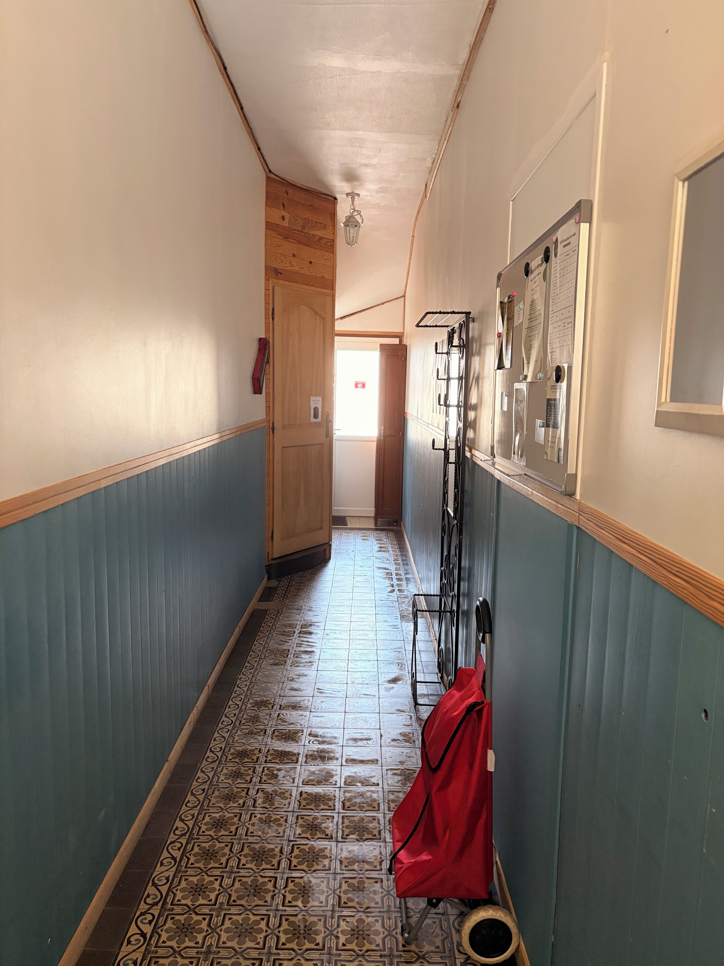 Un couloir étroit, dont le sol est recouvert de carreaux décoratifs, avec des portes en bois et, sur le côté droit, un présentoir métallique sur lequel sont disposés divers objets.