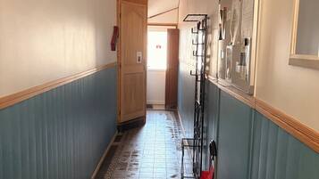 Un couloir étroit, dont le sol est recouvert de carreaux décoratifs, avec des portes en bois et, sur le côté droit, un présentoir métallique sur lequel sont disposés divers objets.