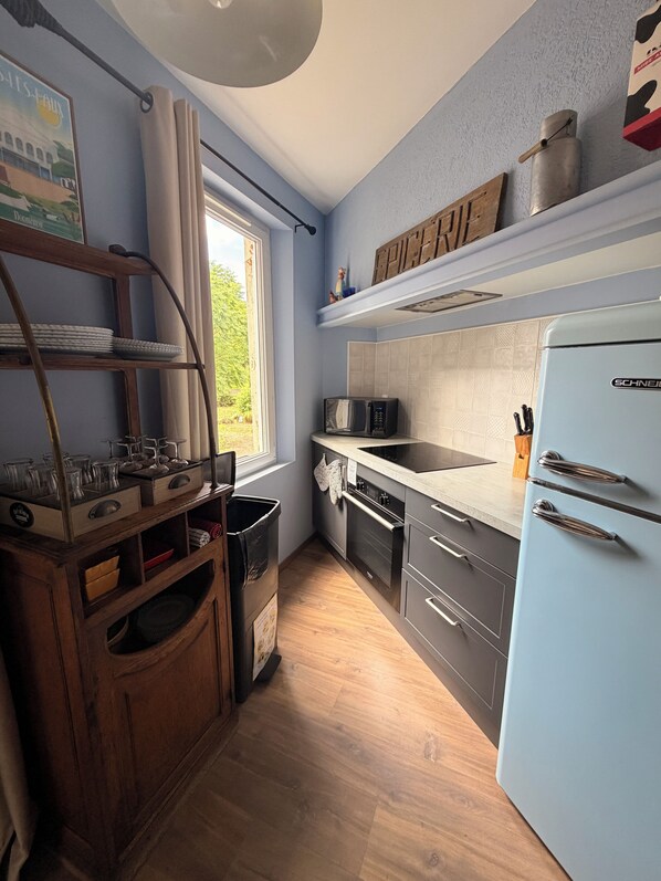 A compact kitchen with a microwave, oven, and a refrigerator. There is a window providing natural light, and a wooden shelf unit with dishes and glasses.