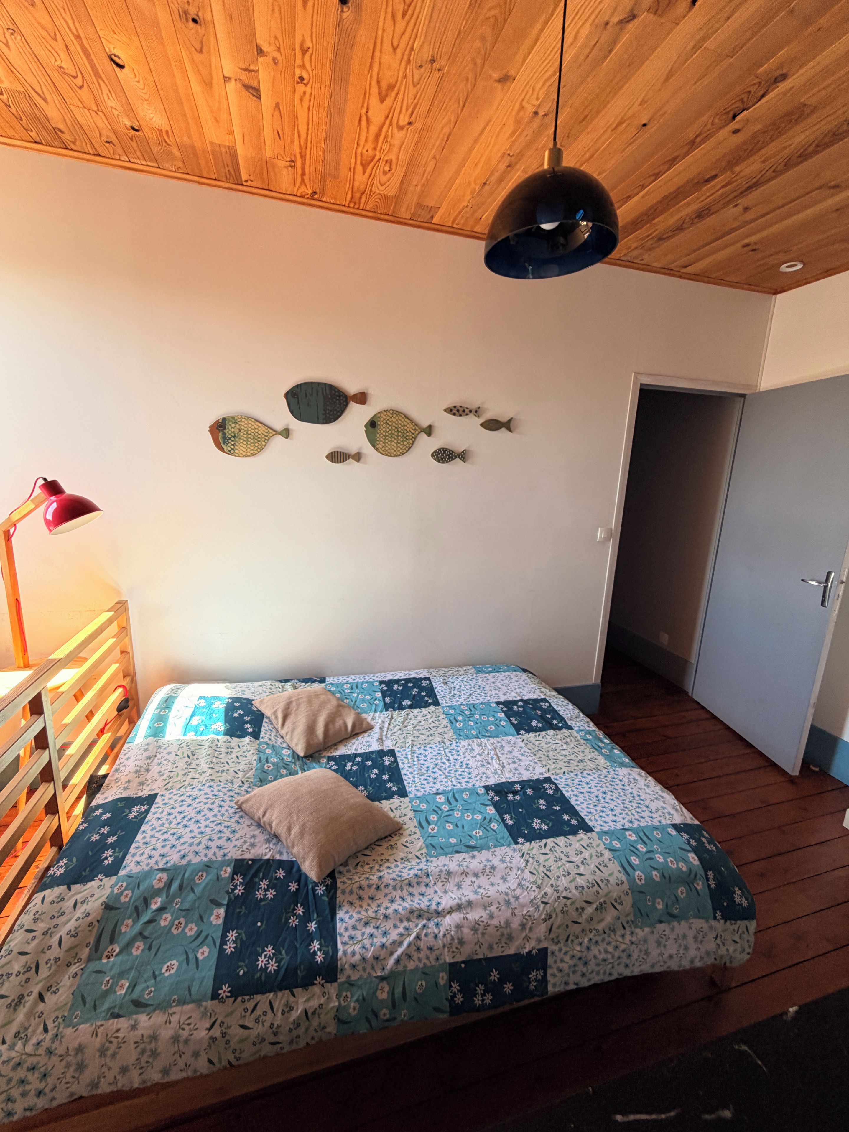A bedroom with a wooden ceiling, a bed with a quilt, a wall-mounted fish decoration, a red lamp, and a door.