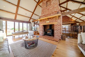A spacious living room with a brick fireplace, wooden beams, a sofa, a coffee table, and large windows. - High Barn - sleeps 8 guests  in 4 bedrooms (Binham)