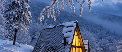 Une cabane enneigée, avec un toit en pente, entourée d’arbres chargés de neige et dominée par un paysage montagneux.