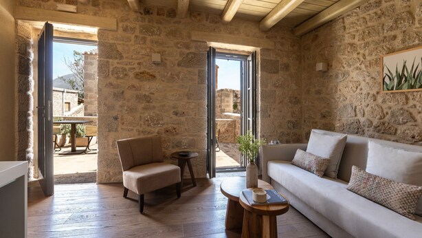 A stone-walled room with a sofa, a chair, a small table, and a door leading outside.