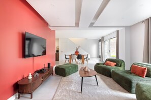 Un salon moderne avec un mur d’accent rouge, un téléviseur à écran plat, des canapés recouverts d’un tissu vert, une console en bois et un coin repas avec une table en verre.