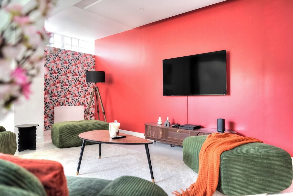Un salon moderne avec un mur d’accent rouge, un téléviseur à écran plat fixé au-dessus d’un meuble en bois et des meubles recouverts d’un tissu vert.