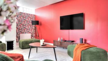 Un salon moderne avec un mur d’accent rouge, un téléviseur à écran plat fixé au-dessus d’un meuble en bois et des meubles recouverts d’un tissu vert.
