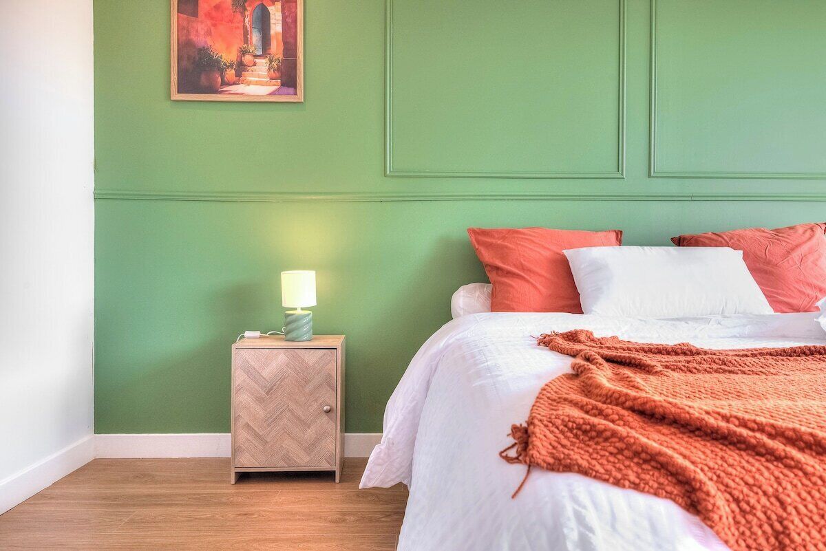 Un dormitorio con cama, almohadas, una mesita de noche con lámpara y un cuadro en la pared.