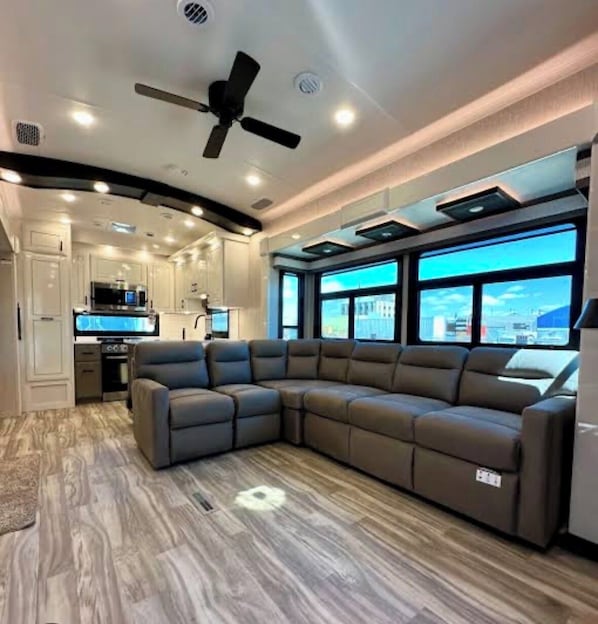 Un espace de vie moderne comprenant un canapé d’angle, un ventilateur de plafond, un éclairage encastré et une vue dégagée grâce à de grandes fenêtres.